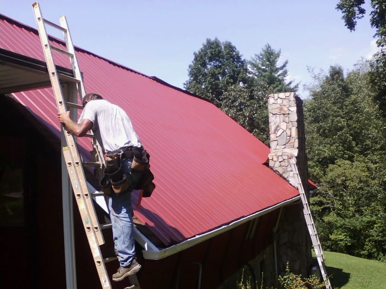 Roofer on a ladder beside a red metal roof and stone chimney — Soffit & Fascia Repair by Roofing Brothers in Ashburn